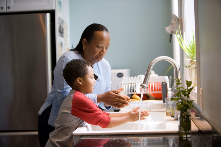 A woman and child are at a kitchen sink. The woman is wearing a blue shirt and dark pants, washing her hands next to a boy wearing a red and white shirt, who is also washing his hands.