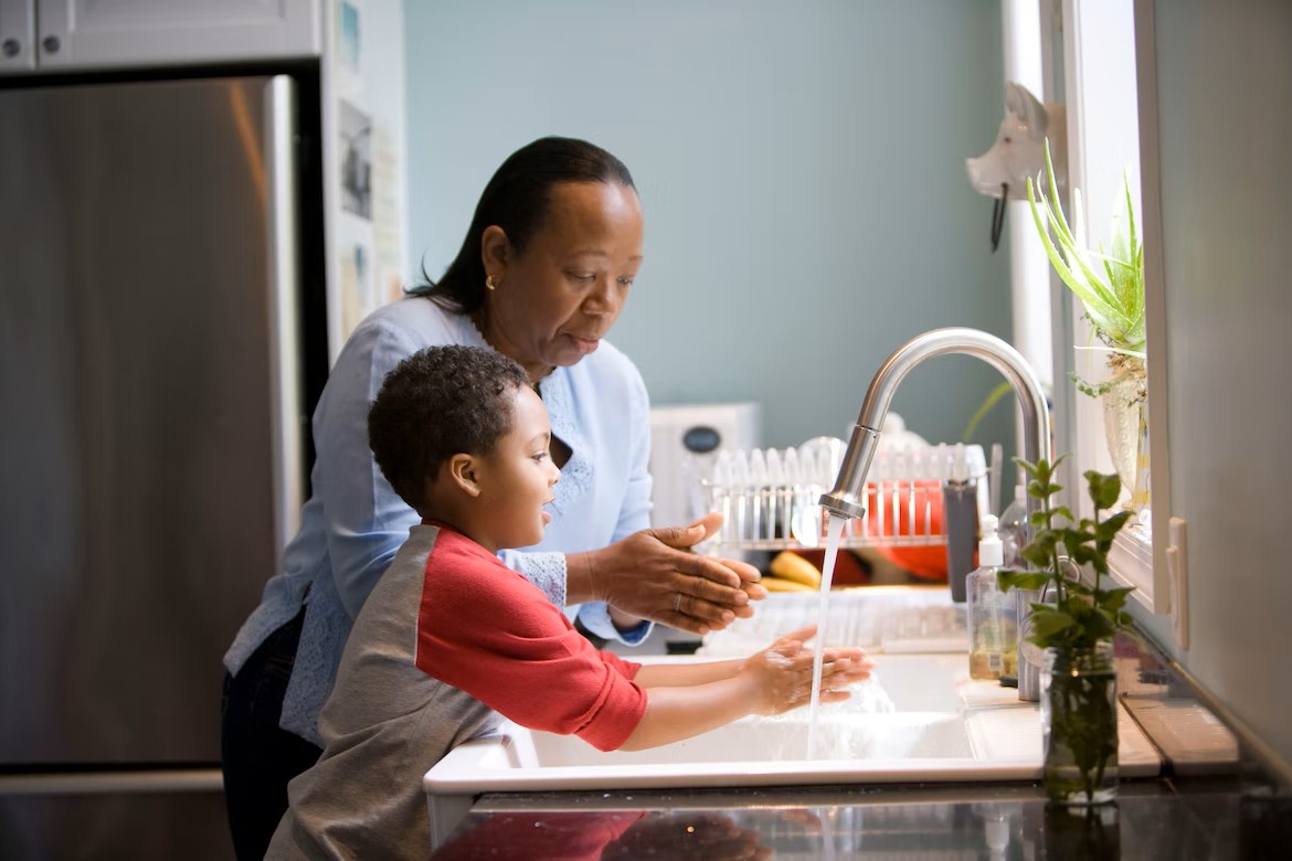 A woman and child are at a kitchen sink. The woman is wearing a blue shirt and dark pants, washing her hands next to a boy wearing a red and white shirt, who is also washing his hands.