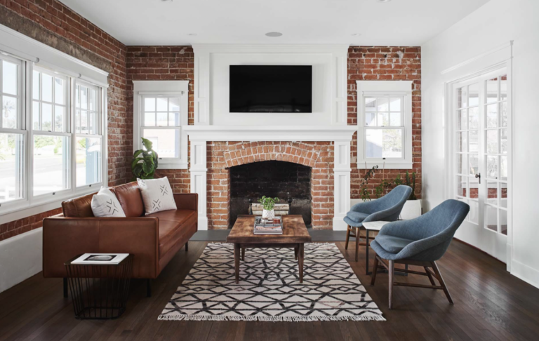 An open living room with wood floors and brick walls features a leather couch, two blue chairs, a patterned rug and a wooden coffee table in the center of the room.