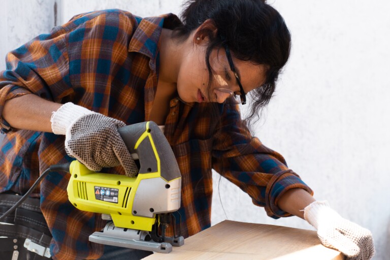 A person is cutting a piece of plywood with a yellow power tool and wearing utility gloves and safety glasses.
