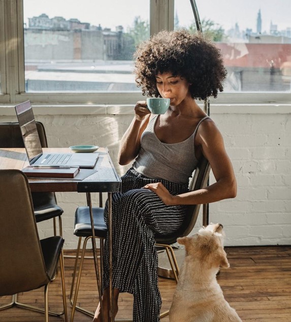 A remote worker is sitting in their home at their kitchen table, drinking from a mug and glancing down at a dog. They are wearing a gray tank top and black lounge pants. 