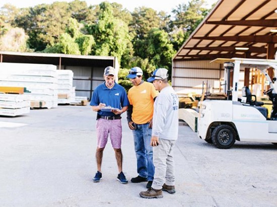 Three Brand Vaughan Lumber employees talking in their lumber yard. There are forklifts and lumber stacks in the background. 