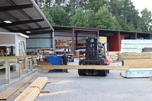 In the image, a forklift is carrying planks of wood in a lumberyard.